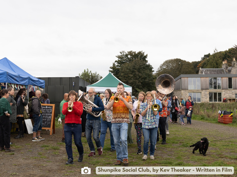 A large brass band (Shunpike Social Club) walk towards the camera past a mix of people and gazebos with stalls