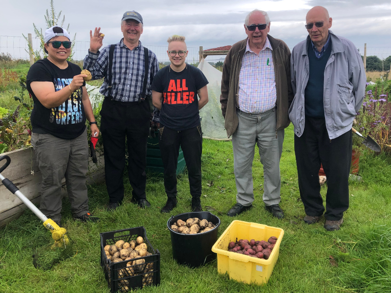 Five people stand by a raised bed in the allotment plot triumphantly showing their potato harvest, which is in boxes at their feet.