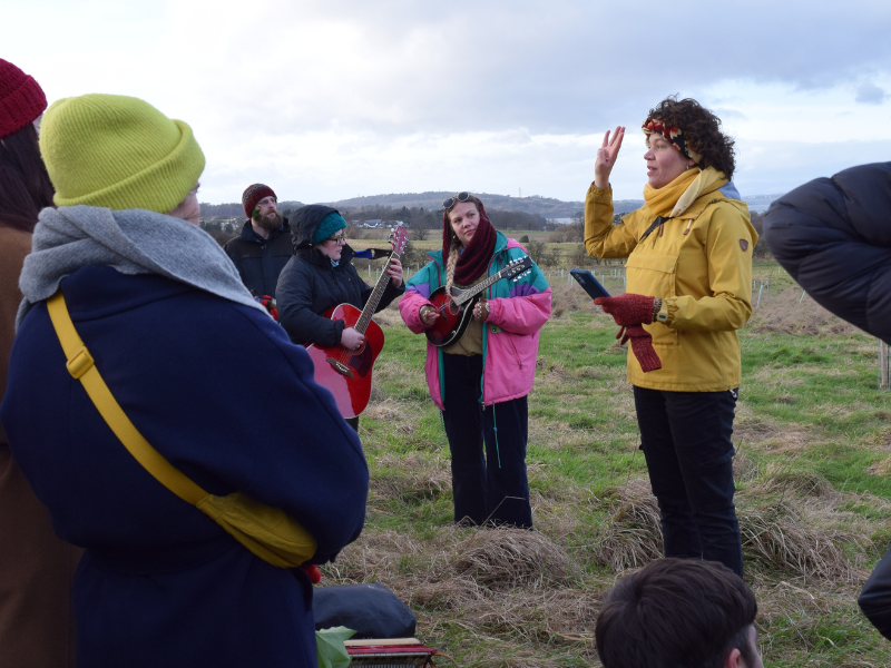 A group of people listen to a song-leader in a yellow jacket. There are two musicians - one with guitar and one with a mandolin, and beyond them, views of the farm fields, with the Firth or Forth and Fife in the background.