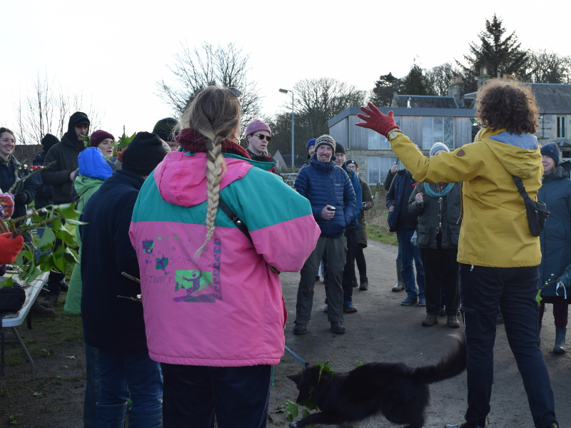 A group gathered at the farm listening to a song-leader in a yellow jacket. Some of the group (and a black dog) are wearing leafy adornments, there are lots of smiling and laughing faces.