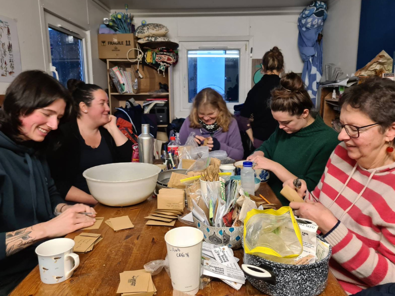 A photo of a group of people in a portacabin sat around a table smiling and working to sort and package seeds into brown envelopes
