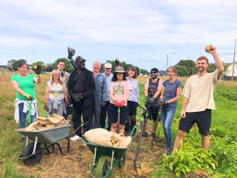 A group of 11 people, all smiling, standing behind two wheelbarrows full of potatoes. Three hold potatoes aloft triumphantly