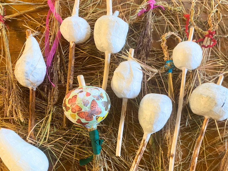 A selection of different vegetable shaped 'seed rattles' made of paper casts with wooden handlies, lying on decorative grasses. One is decorated with illustrations of butterflies