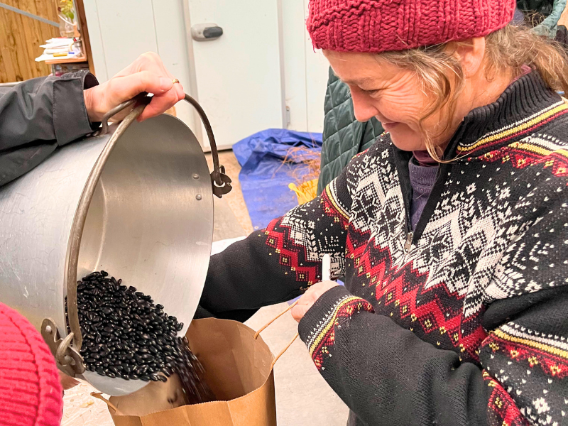 A photo of a smiling member of the farm team. holding open a paper bag as someone else pours in shiny dark seeds from a large bucket