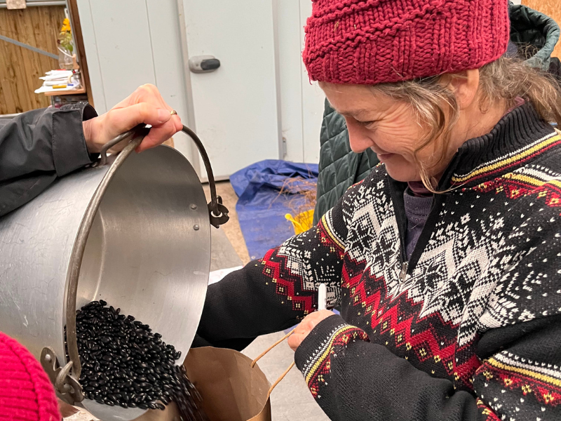 2 A delighted grower in a wooly jumper and hat holds a paper open to collect shiny black seeds being poured from a large pan