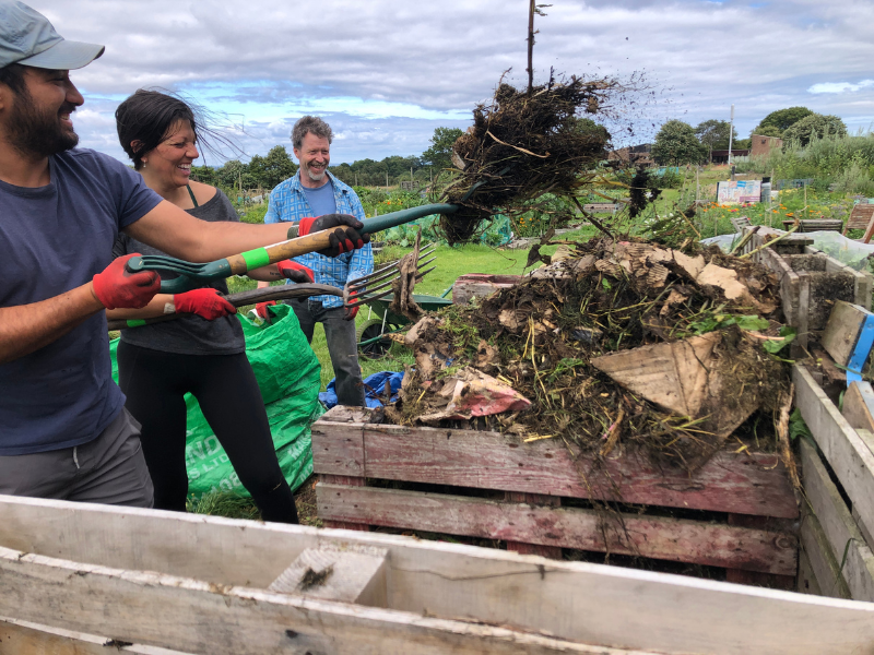 A photo of smiling people forking compost materials into bays in the community allotments