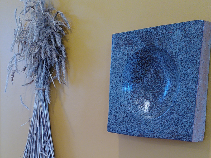 A large, shiny clay 'quern' hanging on a yellow gallery wall, beside a sheaf of wheat