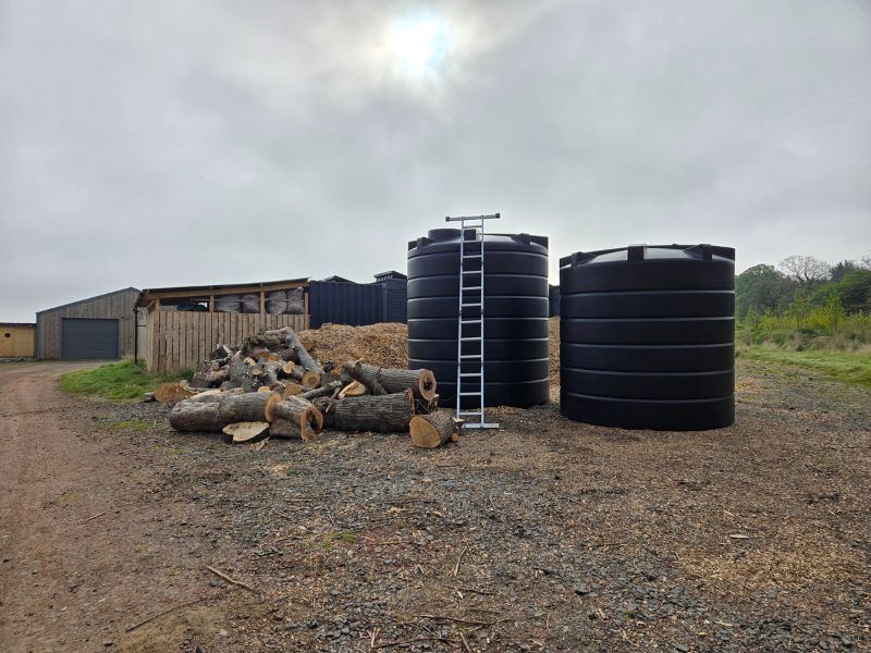 Two enormous black water tanks next to a pile of cut wood and woodchips, with cabins and the pack shed visible behind. One has a ladder leaning on it
