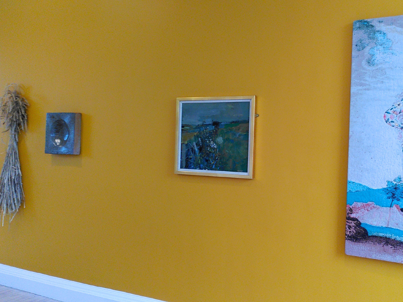 A photo of a yellow gallery wall, with a painting in the middle, a clay quern and sheaf of wheat to the left, and part of the textile piece hanging to the right