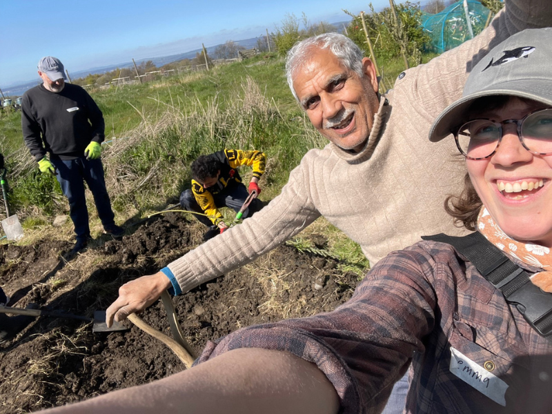 A selfie of two smiling people with muddy hands and tools, and two volunteers behind them working on digging out a pond, in the community garden